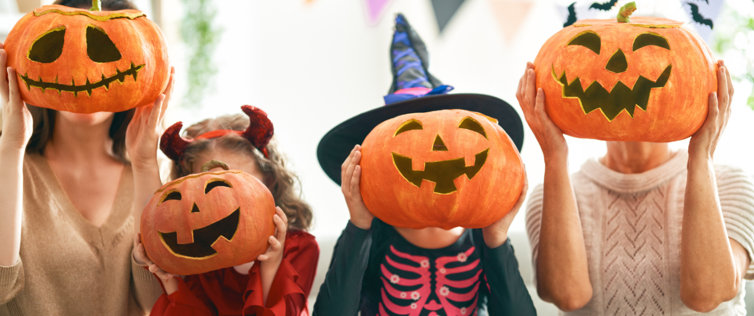 Parents and two young children wearing Halloween outfits, hold pumpkins in front of their faces.