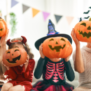 Parents and two young children wearing Halloween outfits, hold pumpkins in front of their faces.