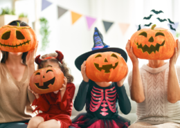 Parents and two young children wearing Halloween outfits, hold pumpkins in front of their faces.